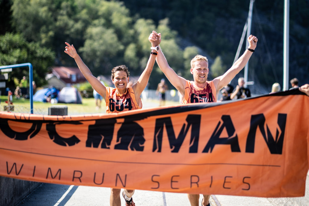 A mixed pair of competitors reach the finish banner of the Rockman swimrun event in Norway, running with arms in the air.