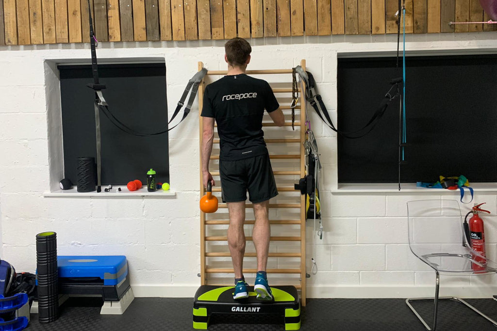 A male runner stood on the edge of a step doing weighted single leg calf raises holding an orange kettlebell