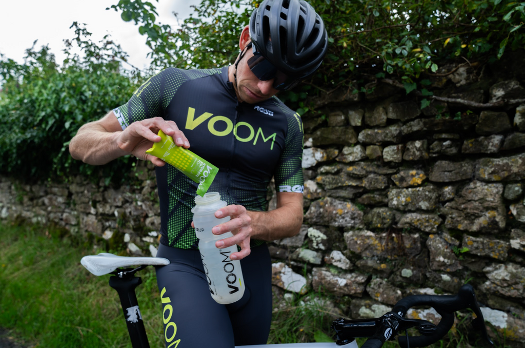 A male cyclists in VOOM Nutrition team kit standing over his bicycle tipping a sachet of hydration powder into a water bottle