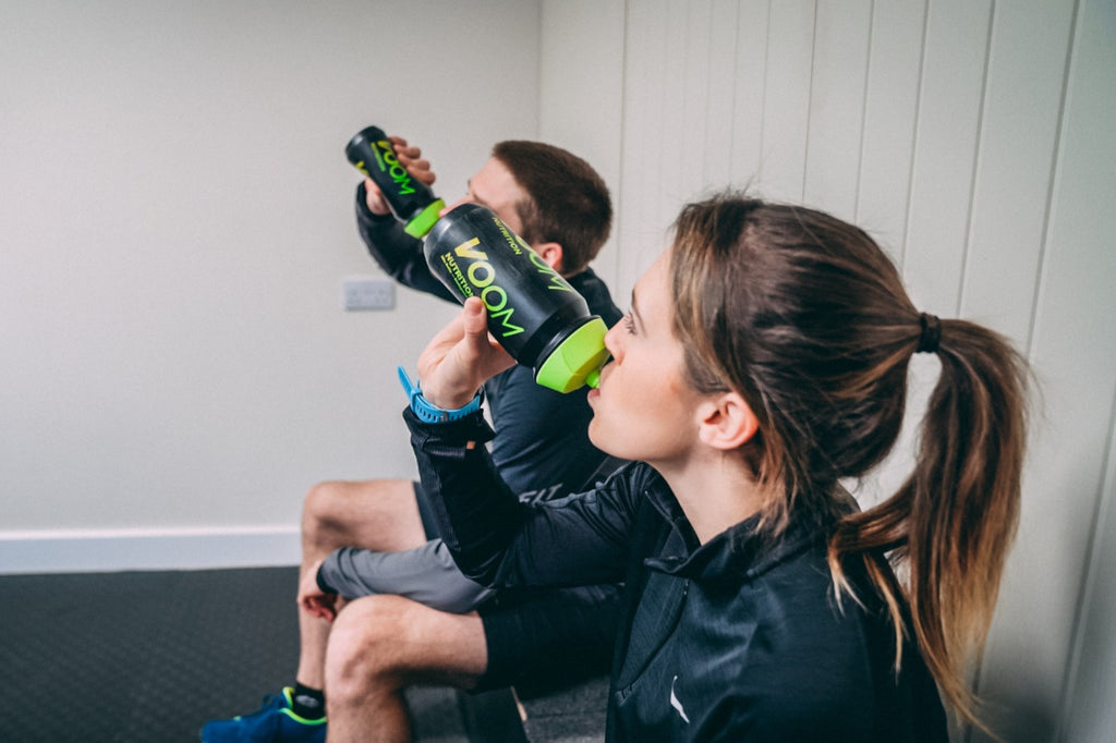A male and female athlete sat drinking VOOM Hydrate from a black and green VOOM bottle whilst on a rest period during a gym session