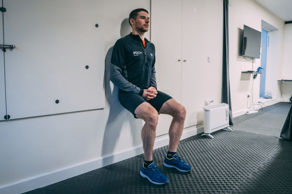 A man squats against a wall holding a wall sit position in the gym