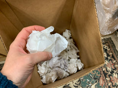 A hand crumpling used label paper backing and placing it in a bin of crumpled paper for re-use as void fill in shipping boxes