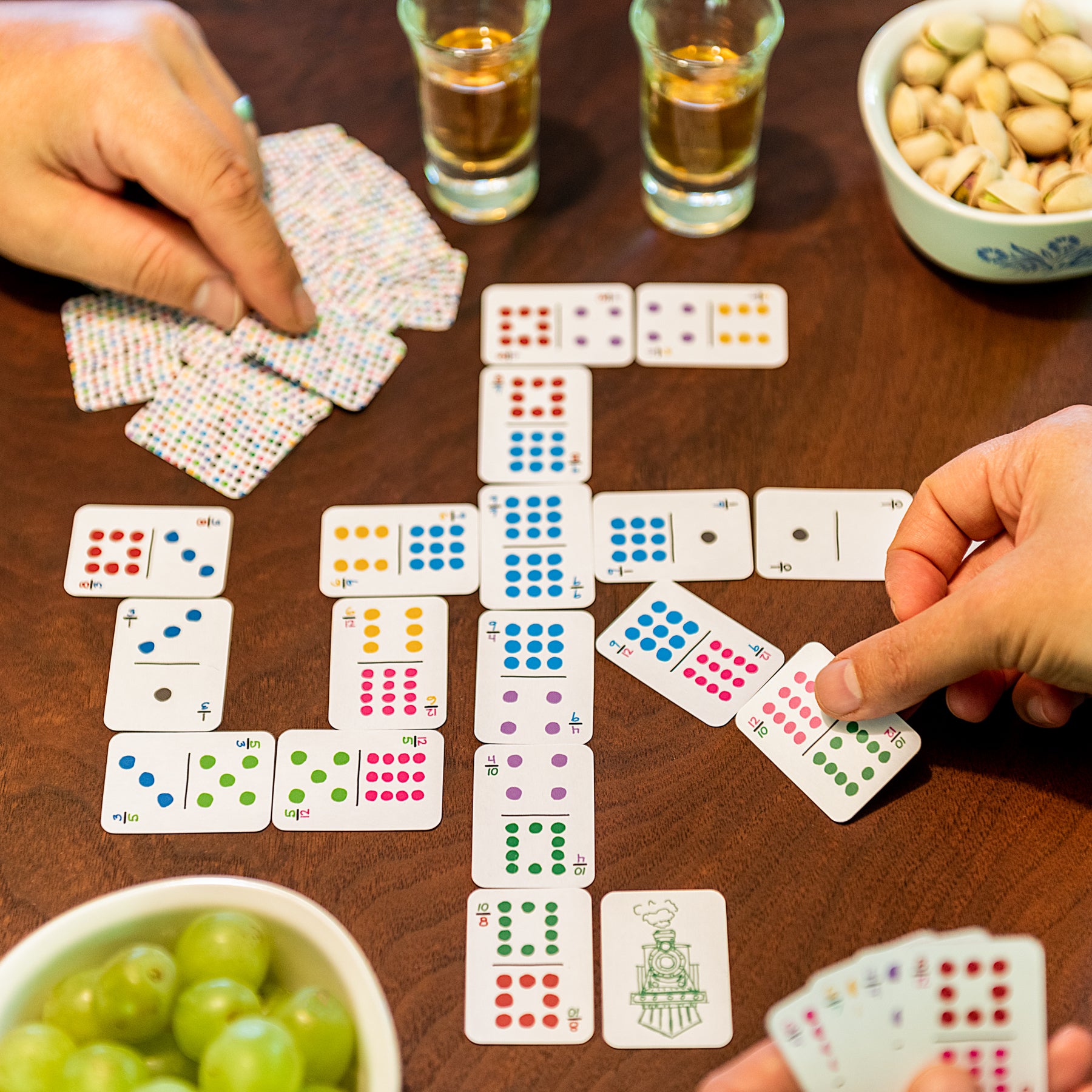 Two people playing Mexican Train dominoes with colorful small playing card dominoes instead of tiles. The game takes up less space on the table for a travel size. Strewn on the table are snacks and drinks from a game night.