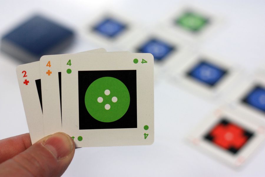 A hand holding up two tiny square cards from the game IOTA. The card has a number, color, and shape, and each hard has a unique combination of those 3 elements.