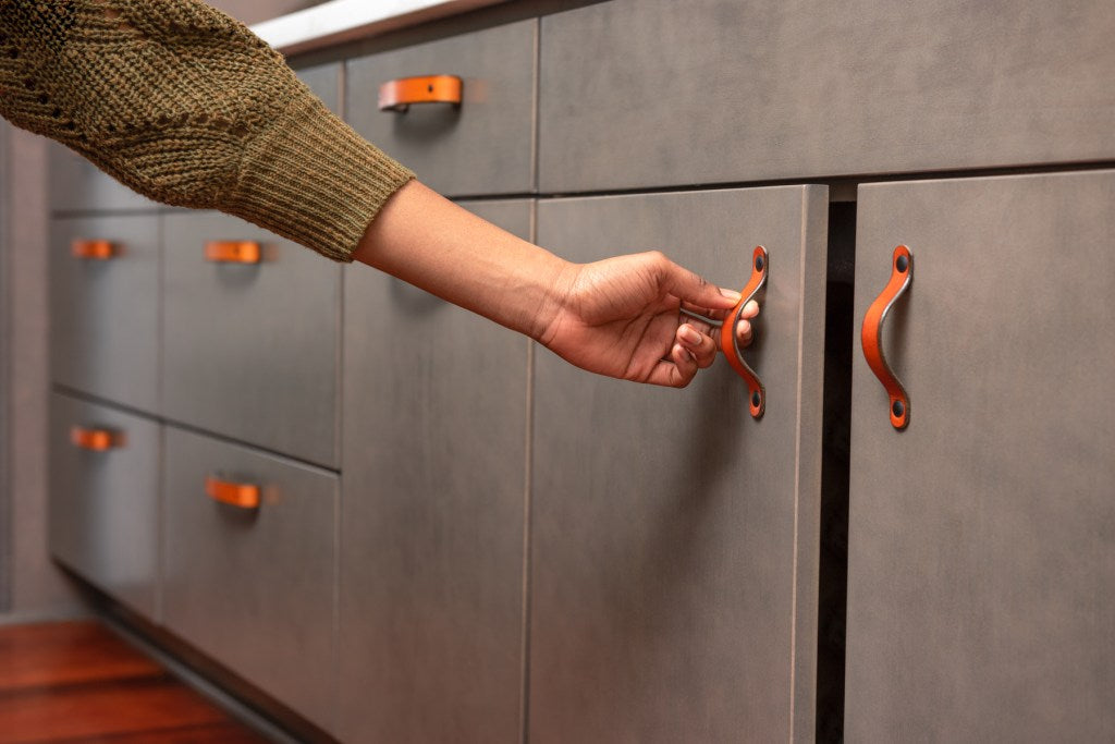 Black woman wearing an olive sweater reaching for a honey leather cabinet door handle underneath a kitchen sink. 