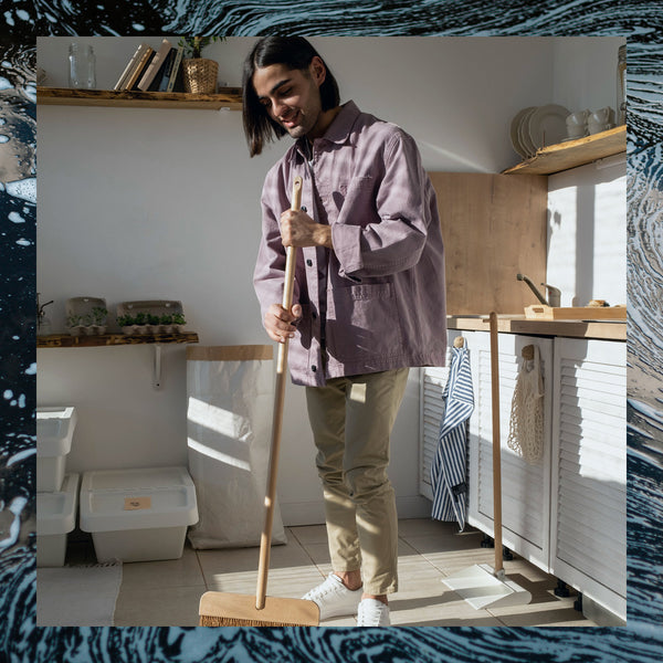 A long haired man holding a broom in a kitchen lit by afternoon sun