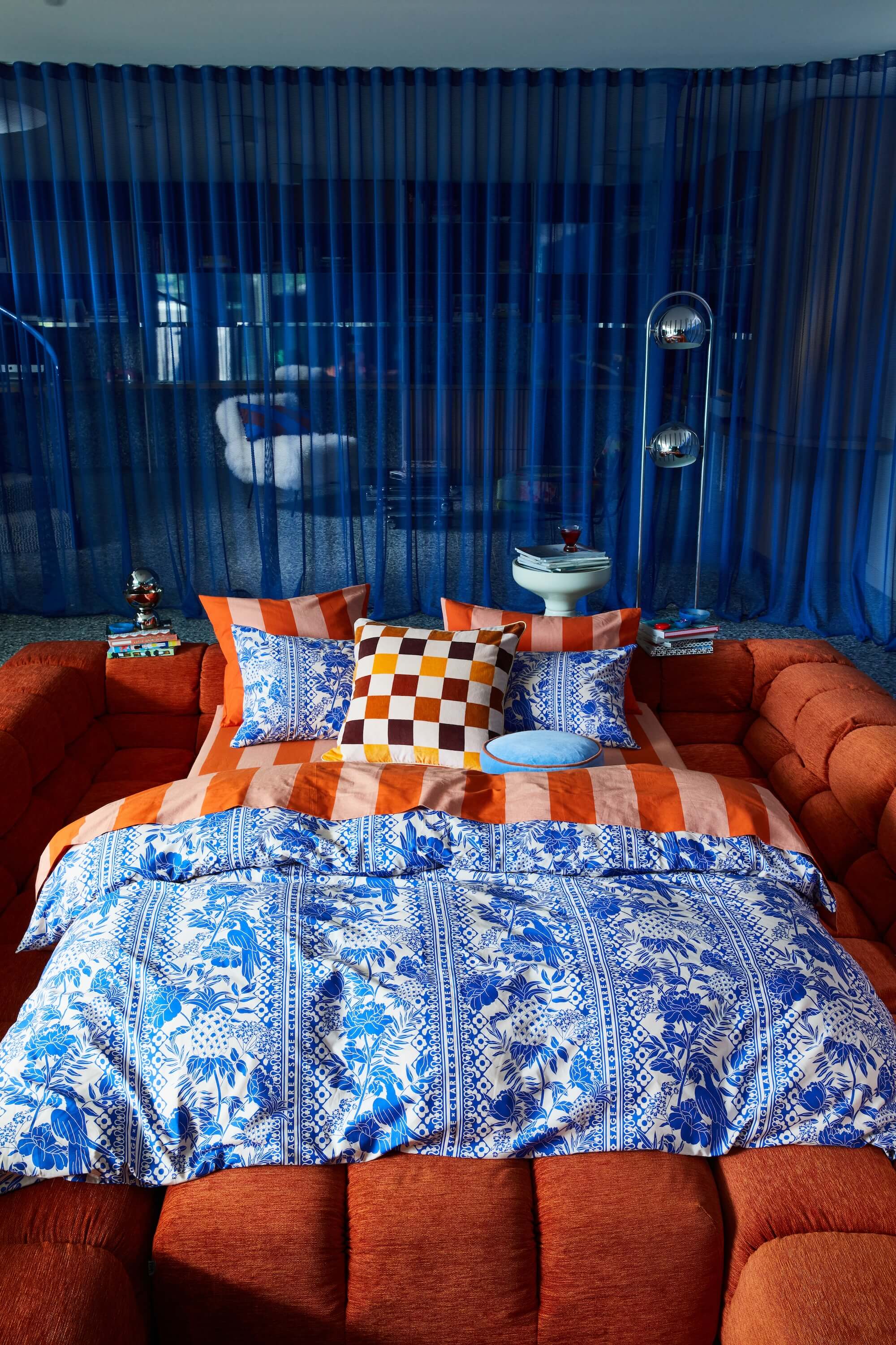 blue and orange bedding in front of blue sheer curtain and orange sofa