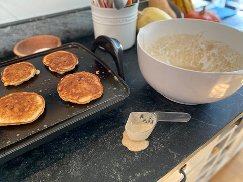 image of a skillet on a counter with pancakes cooking and a white mixing bowl in the background with a measuring cup
