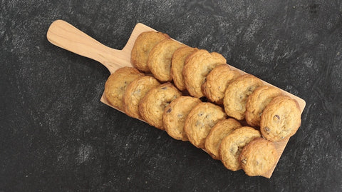 two rows of chocolate chip cookies on a wooden peel on a dark countertop