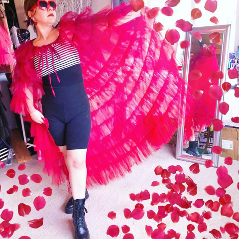 A woman with a long and ruffled tulle red cape with rose petals spreading from out of her cape. She is wearing heart shaped glasses, black shorts and boots, and a black and white stripped top.
