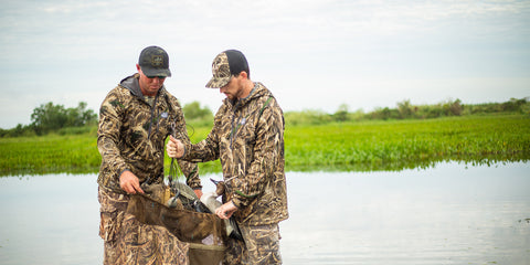 Two duck hunters play decoys in bag in front of a body of water. They are wearing Sportsman Gear Outbound Hoodies.