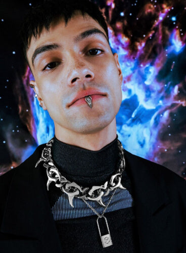 Man with piercings and necklaces against a cosmic background.