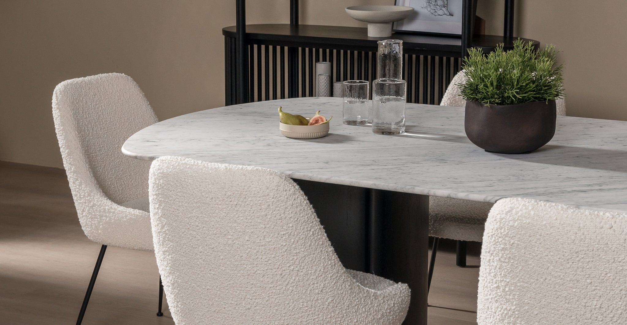 Tathra Marble Dining table from The Loom Collection shop with black and white boucle dining chairs and a black shelf in the background. The dining table has a plant on the White marble top and a carafe with glasses and fruit for styling. 
