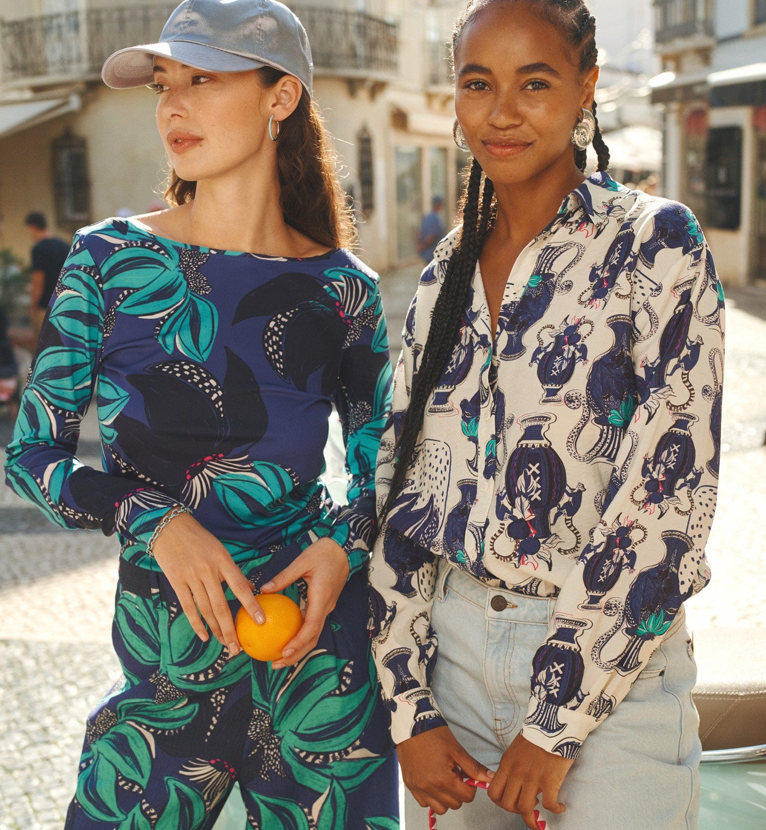 Two women in patterned outfits, standing outdoors in a sunny urban setting.