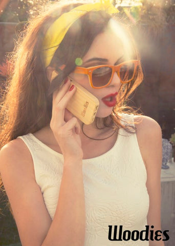 girl in white sleeveless top wearing mustard shade of headband and solid orange sunglasses talking on the phone with a wooden casing