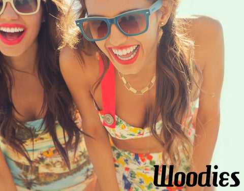 2 girls in floral bikini wearing light brown and blue wooden sunglasses laughing