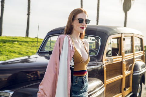 girl in olive top holding a pink and white jacket wearing a wooden sunglasses in the shade of black sitting on the hood of a wagon