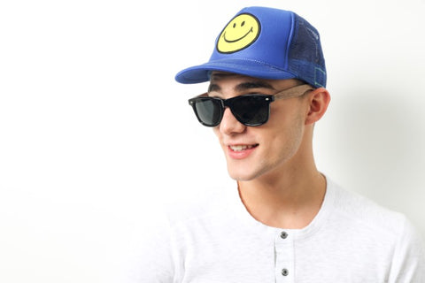 guy in white round neck long sleeves shirt wearing a blue cap with yellow smiley in the middle wearing a wooden sunglasses