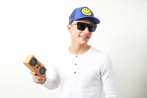 guy in white round neck long sleeves shirt wearing a blue cap with yellow smiley in the middle wearing a wooden sunglasses holding a wooden speaker