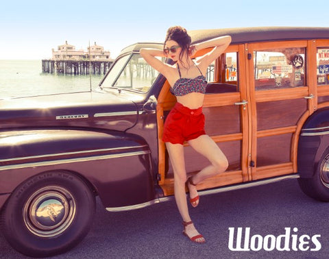 girl in floral top and red short and red sandal wearing wooden brown sunglasses beside a vintage wagon