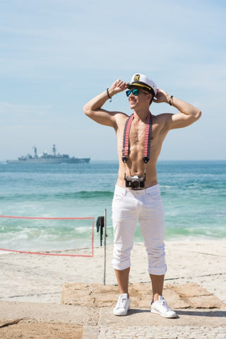 topless guy in captain's hat wearing wooden sunglasses in the shade of bluish green with camera hanging from his neck