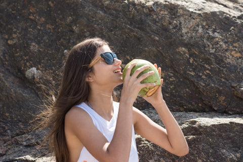 girl with long brown hair wearing wooden sunglasses in the shade of black drinking from a coconut