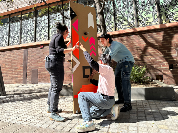 The adults stand around a cardboard pillar. A woman in black touches a pattern on the cardboard and with her other hand her finger connects to the middle adult who touches a different pattern on the cardboard. The third participant holds the hand of the middle participant and touches the earth pattern on the other side of the cardboard pillar.
