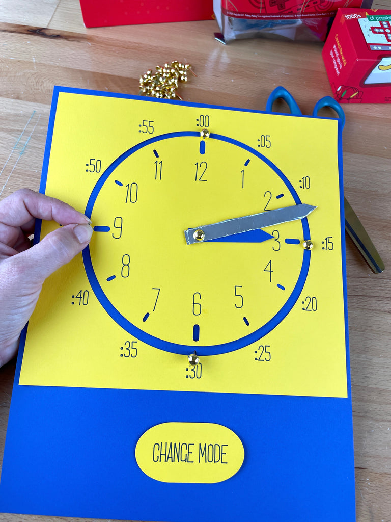 Hand placing brads on 12,3,6,9 on a paper clock on yellow cardstock