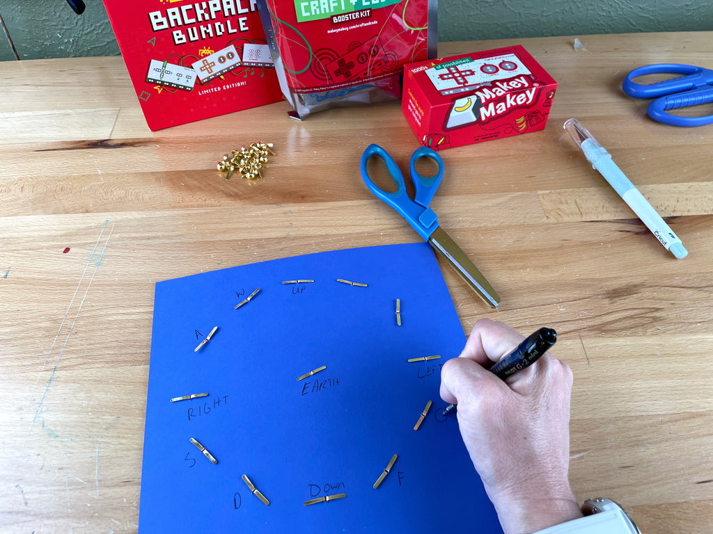 Blue cardstock (back of the clock) there are brass fasteners placed at all the five minute increments. A hand is labeling each point with the Makey Makey key press for each spot.