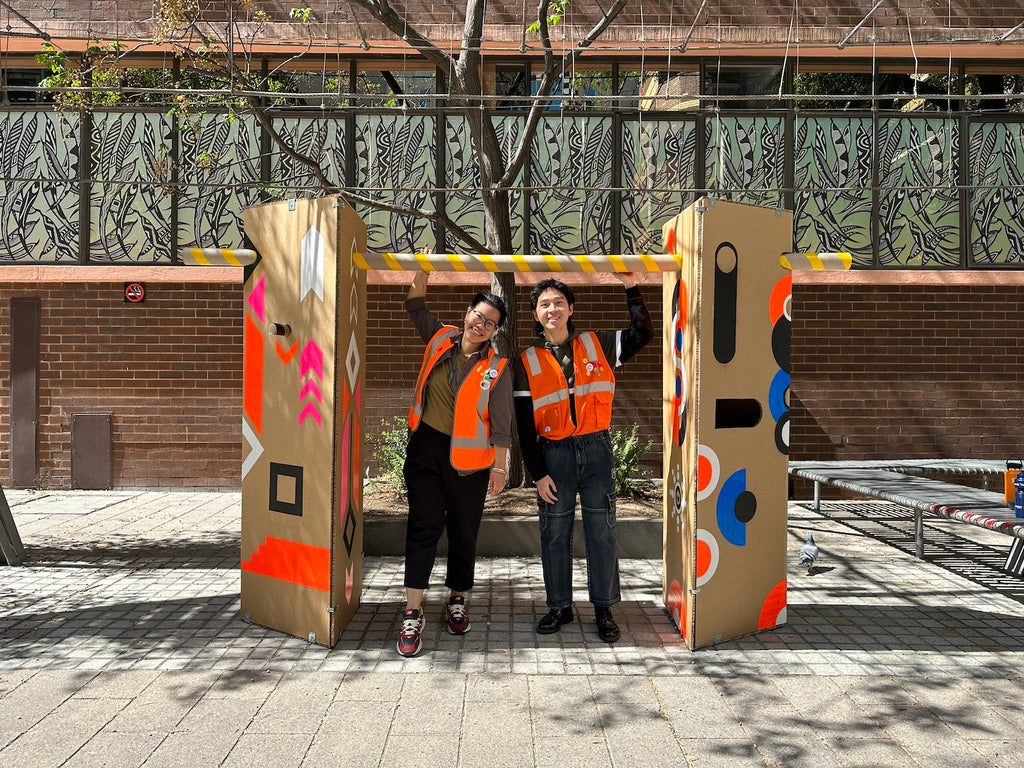 Two cardboard pillars covered with geometric shapes. A woman and man stand side by side in between the pillars holding onto the tube that connects the pillars.