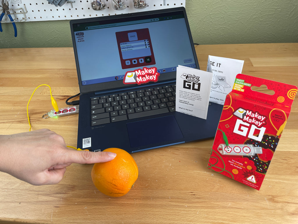 Makey Go Packaging in the foreground and a hand touching an orange