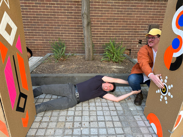 There are two cardboard pillars with colorful shapes. A man in a yellow hat is squatting to put his hand on the power pattern on the pillar on the right. Another man lays on the ground in effort to make a connection across the pillars, he has his feet touching the second pillar and is grabbing the leg of the man in the yellow hat.