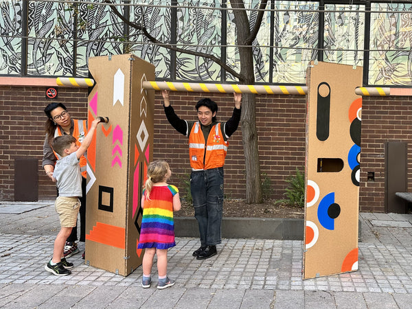 The full installation of Symphony With is shown with two large cardboard triangular pillars and a connecting cardboard tube. A man in an orange vest is holding onto the tube and talking to a small child in a rainbow dress that is looking at the exhibit.