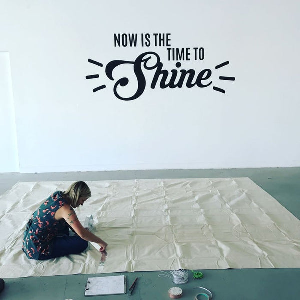 Woman works on large mat to create a maze. The wall behind her says "Now is the time to shine"