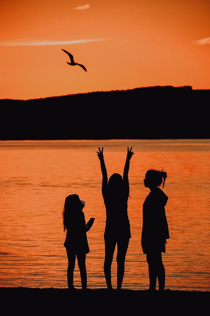 silhouette photo of 3 kids in front of lake with orange colored sky