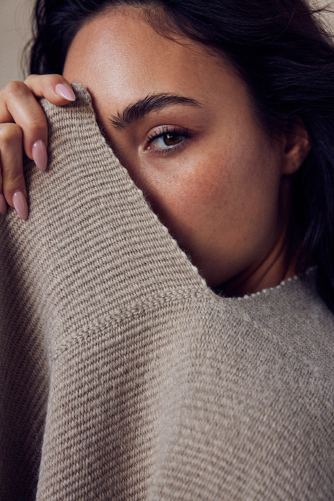 Close-up of a woman partially hiding her face behind a beige sweater, showcasing her natural beauty.