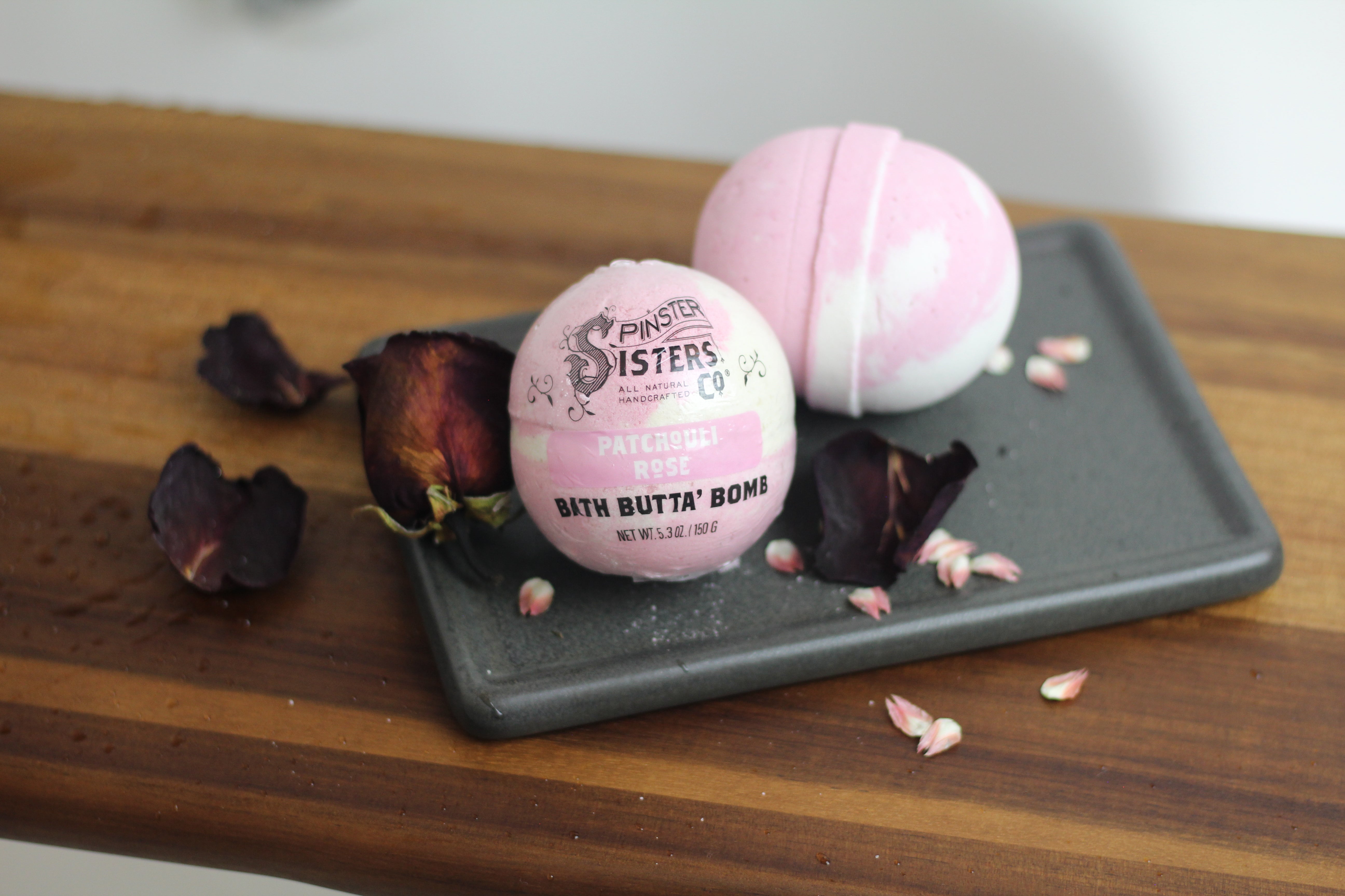 Two Patchouli Rose Bath Butta' Bombs surrounded by dried rose petals and patchouli on a bath tray