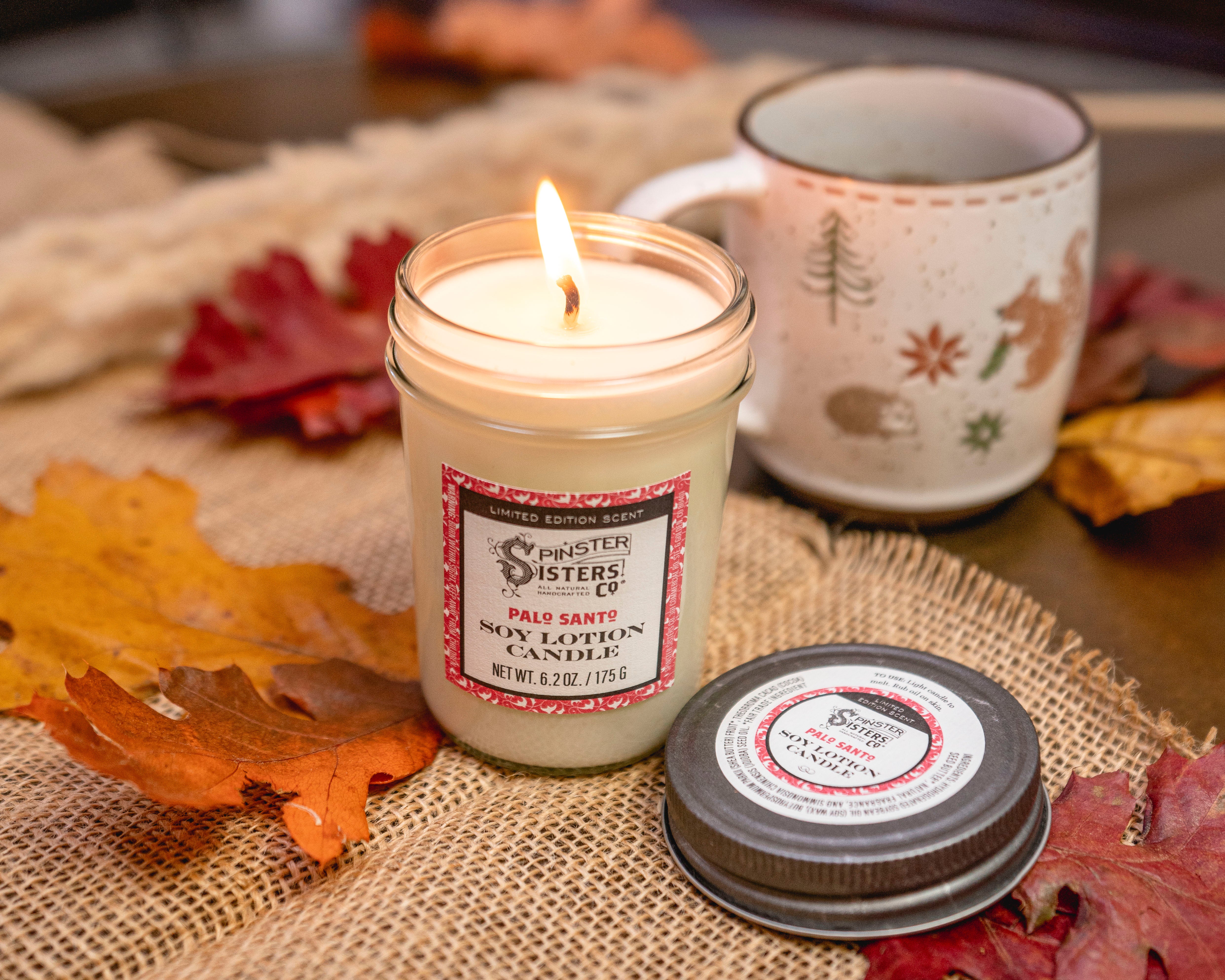 A Palo Santo scented Soy Lotion Candle burns in its jar next to its lid, a coffee mug and among scattered autumn leaves