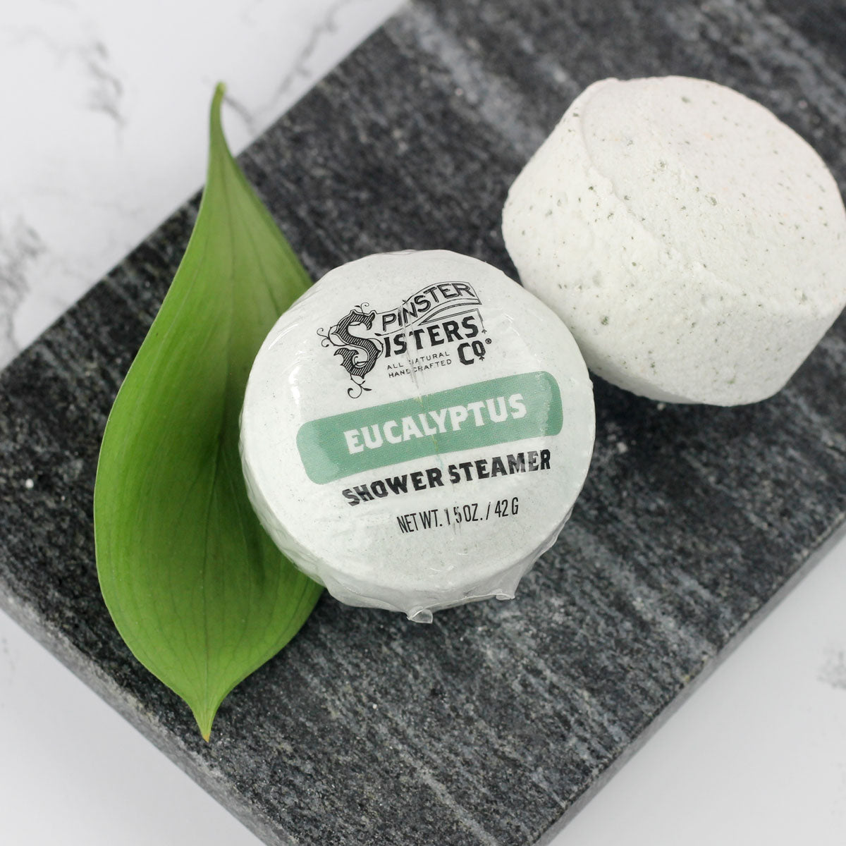 pale green eucalyptus shower steamer pellet wrapped in bioplastic on gray stone next to green leaf