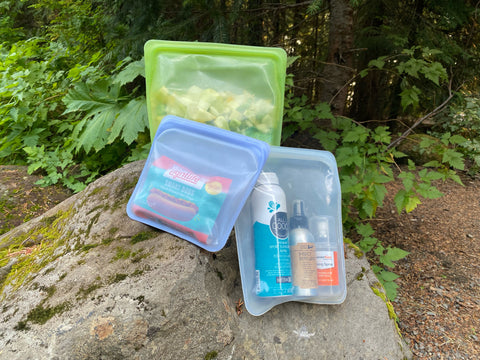 Three silicone Stasher Bags of various sizes are resting on a rock. The largest has chopped veggies in it, the second largest has sunscreen and bug spray, and the smallest is a purple sandwich bag size and has a package of plant-based hot dogs in it.