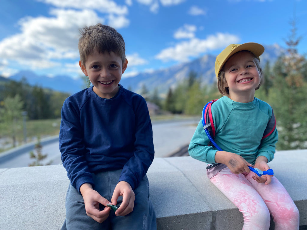 photo of two kids sitting on rock with mountains and forest in the background