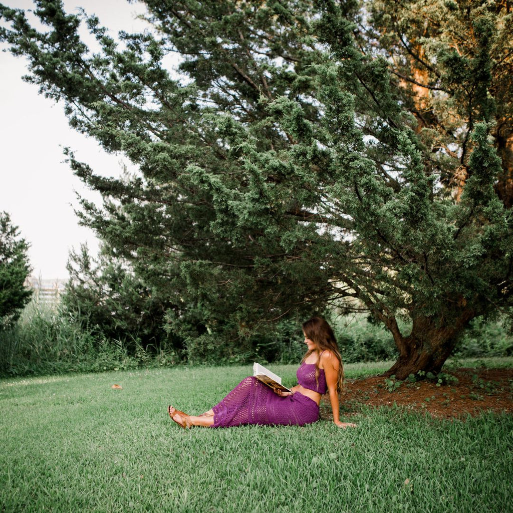 A woman sits in a field reading a book, she's wearing a crocheted outfit consisting of a matching purple cropped tank and maxi skirt