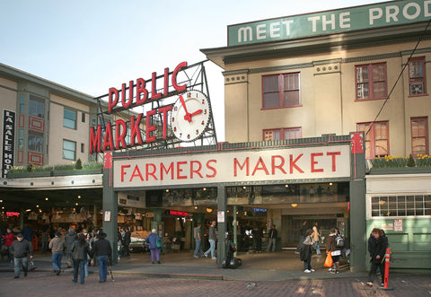 Pike Place Market's main entrance. A red neon sign says Public Market over another sign with red letters that says Farmers Market. Several people walk in front of the signs on a brick street. 