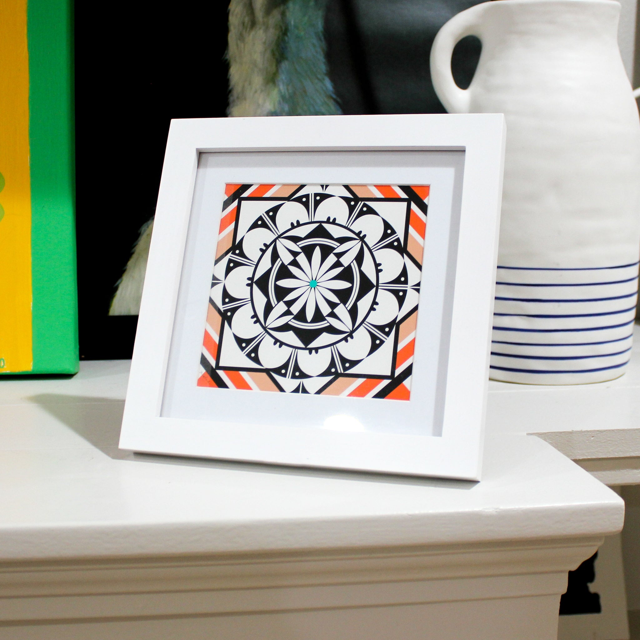 A-black-white-and-orange/red-geometric-design-with-a-flower-in-the-center-sits-on-a-shelf-in-a-white-frame