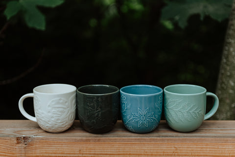 Four Native-designed mugs in white, black, blue, and green, sit on a wooden rail