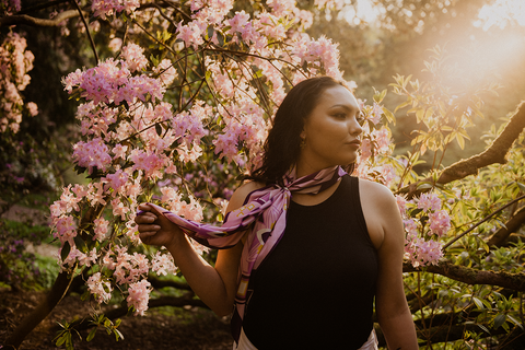 A woman wearing a black tank top and a purple neck scarf stands in front of purple flowers. The lighting makes the photo look like a Renaissance painting.