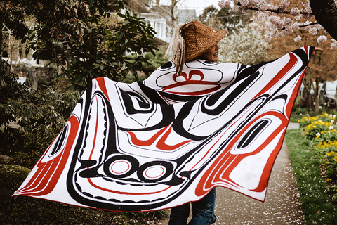 A woman in a traditional Salish cedar hat twirls in a black, white, and red blanket featuring Northwest Coast formline design