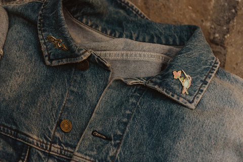A denim jacket lapel, featuring an enamel pin with a hummingbird sitting on an elderberry branch