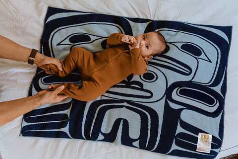 A sweet Native baby in a brown onesie rests on a blue baby blanket that features Northwest Coast designs