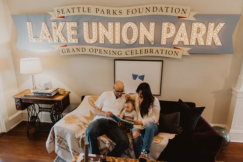 A family of three (dad, toddler, and mom) sit on a couch in a nice living room. A vintage sign above them says LAKE UNION PARK.